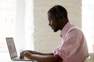Happy African American student wearing headphones taking online studying course, reading learning materials at laptop, typing important educational information, e-learning person